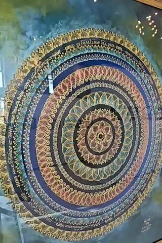 This is a close-up, panning video of a circular mandala artwork. The artwork is intricate and colorful, featuring concentric circles of decorative patterns. The dominant colors are shades of blue, gold, teal, and pink. There are reflections on the surface of the artwork, suggesting it is behind glass or a similar protective layer. The lighting is somewhat dim, and the background is dark. The video pans slowly from the top to the bottom of the artwork, showcasing its detailed design.