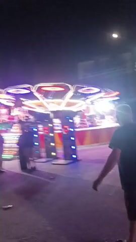 The video opens with a low-angle, shaky shot of a dimly lit outdoor scene at night. A young man in a black t-shirt and black shorts is the main subject, walking away from the camera and towards a brightly lit amusement ride in the background. The ride appears to be a spinning attraction with multiple lights and platforms. Several other people are visible near the ride, some standing and others appearing to be on the ride itself. The ground is wet and reflective, suggesting recent rain or puddles. The overall atmosphere is that of a carnival or fairground at night, with the moon faintly visible in the dark sky. The camera movement is unsteady throughout the brief clip.