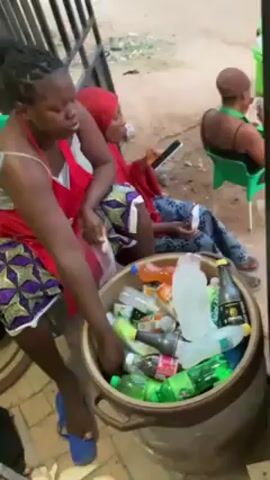 The video shows a street scene with several people.  Three women are sitting and relaxing; one is holding a tablet. A woman wearing a red apron and patterned skirt is putting something into a large container full of empty plastic bottles and cans of soda and other beverages.  A man wearing a green-and-white striped shirt is partially visible standing behind a black metal gate. The setting appears to be outdoors, possibly near a street-side shop or gathering area.