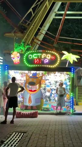 This video captures a nighttime scene at an amusement park or fair. In the foreground, a paved area with drain grates is visible. Several people are present, including a man and a woman standing in front of a game booth. The booth is illuminated with neon lights and features a large display of plush toys. A prominent sign above the booth reads "ОСТРОВ ИГР" (Island of Games) in colorful, blinking letters. A large, cartoon-like cat character in a shirt and tie is prominently displayed within the booth. In the background, a Ferris wheel or similar amusement ride is visible, also illuminated. The overall atmosphere is festive and lively.