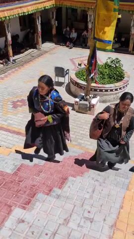 Two women in traditional clothing are walking up a staircase. The woman on the left is wearing a black dress with a blue and embroidered vest, and a brown purse is slung over her shoulder. The woman on the right is wearing a dark dress, a brown vest, and carrying a brown bag on her shoulder. The staircase has red and white patterned steps. In the background, there is a courtyard with people sitting and a building with colorful banners. The scene appears to be in a monastery or a similar cultural site.