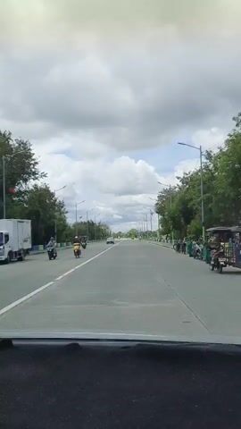 A video shows a view from inside a vehicle, traveling on a paved road with multiple lanes in each direction. The road is lined with trees and streetlights. There are several motorcycles and cars visible on the road. The sky is cloudy.