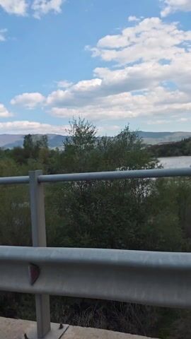The video shows a scenic view from a bridge, possibly taken from a vehicle driving across it. A wide river flows beneath, surrounded by lush greenery and distant mountains under a blue sky with scattered white clouds. The bridge's metal railing is visible in the foreground, offering a partial view of the landscape.