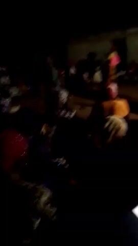 The video is shot at night, in a dimly lit outdoor setting with a group of people. The subjects are scattered around, some sitting and some standing. The quality of the video is poor, making it difficult to discern fine details. Individuals appear to be wearing various articles of clothing, including head coverings. A red helmet is visible in the foreground.