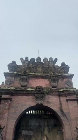 The camera looks up at a brick archway with stone carvings, including statues of winged figures. The archway is old and weathered, with some green moss growing on the surface. Below the arch is a sign that says "ARTIKA'S EMAS DAN PERAK, BATIK DAN LUKISAN, GOLD AND SILVERWORKS, BATIK AND PAINTINGS". The sky is overcast and gray. Two carved figures stand guard on either side of the archway.