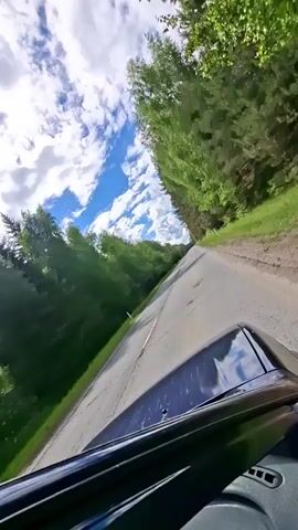 A low-angle, front-facing view captures a vehicle driving along an asphalt road in Lukintsy, Russia. The dark hood of the vehicle is visible at the bottom of the frame, reflecting the bright sky. The road stretches into the distance, flanked on both sides by dense green trees and vegetation, suggesting a rural or forested environment. The sky overhead features patches of blue amidst scattered white cumulus clouds, indicating a clear and sunny day. The road surface shows signs of wear. The camera is slightly tilted to the left, providing a dynamic perspective of the journey. No people or visible text are present in the frame.