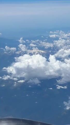 An aerial view from an airplane window shows a landscape below with mountains and clouds. The mountains in the distance appear to be snow-capped, with patches of white snow visible. Fluffy white clouds are scattered across the scene, some obscuring parts of the mountains and the terrain below. The sky above is a gradient of blue, becoming lighter towards the horizon. In the foreground, a portion of the airplane's engine nacelle is visible at the bottom of the frame. The overall impression is of a clear, sunny day with a vast expanse of sky and land.
