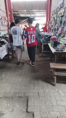 The video shows a busy market stall with many items for sale. In the foreground, a person wearing a red shirt with "SPAIN 10" on the back is walking away from the camera. They are wearing black pants and white sandals. To their left, another person in a white t-shirt is partially visible. The stall itself is filled with various items, including many pairs of flip-flops and sandals displayed on tables and hanging on a wall. There are also some boxes and other miscellaneous goods. Several people are browsing the merchandise. The environment appears to be an open-air market with a corrugated metal roof. The ground is paved with bricks. The lighting suggests it is daytime.