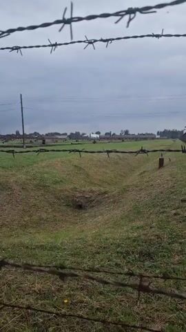 The video shows a view through barbed wire fences. In the foreground, barbed wire is visible, with a grassy field behind it. There are some depressions in the field that may be trenches. In the background, several long, low buildings are visible, arranged in rows. There are also some trees and utility poles. The sky is overcast. The overall impression is of a somber and desolate scene.