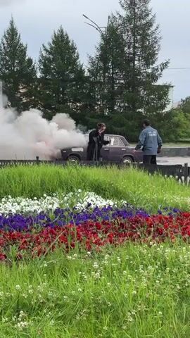 A dark-colored car is engulfed in smoke and flames, positioned next to a flowerbed. Two individuals are present near the car. One individual, wearing dark clothing, appears to be interacting with the car or the smoke. The other individual, wearing a light blue jacket with a darker collar and dark pants, stands nearby, observing the scene. The car is on a paved surface, and a grassy area with a flowerbed in front separates it from the viewer. Trees and buildings are visible in the background. The flowerbed contains flowers in red, white, and purple, arranged in horizontal bands. The sky is overcast.