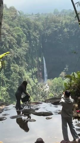 The video shows two people standing on a rocky ledge overlooking a waterfall. The person on the left is a woman wearing gray sweatpants and a dark top. She is standing next to a black folding chair and appears to be adjusting it. The person on the right is a man wearing a gray hoodie and gray sweatpants. He is holding a phone and taking a picture of the waterfall. The waterfall is tall and thin, surrounded by lush green trees. There is a pool of water in the foreground, reflecting the people and the scenery. The weather appears to be sunny and clear.