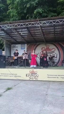 A band performs on an outdoor stage. There are six people on stage. On the far left, a man plays an electric guitar. Next to him, a man plays an accordion. In the center, a woman in a red dress sings into a microphone. To her right, a woman in a black dress also sings into a microphone. Behind them, a man plays a bass guitar. On the far right, a man plays a drum set. The stage has a metal frame and a roof. Behind the band, there are two large banners, one with a QR code and text, and another with a circular graphic and text. In front of the stage, there's a long banner with a logo and text. The ground in front of the stage is concrete. In the background, there are many green trees.