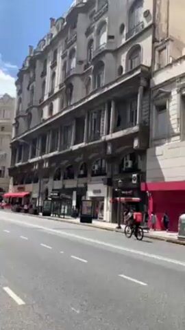 The video shows a street scene in a city with European-style architecture. The camera is moving, likely from a vehicle on the road. On the right side of the road is a tall, ornate building with multiple floors and columns. There are shops with red awnings at street level. People are walking on the sidewalk. A cyclist wearing red is riding on the street. A bus passes by in the distance on the left side of the road. The sky is blue with some clouds.