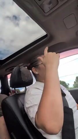A person is driving a car with the sunroof open. They are wearing glasses and a white t-shirt. They have their arm raised to their head. The person appears to be singing or talking. The interior of the car is visible, as well as the sky and trees through the open sunroof and window.