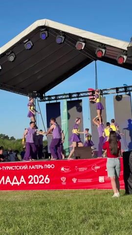 A medium shot of a stage set up outdoors, on which a group of people is performing. The group consists of about twelve people, most of whom are wearing purple pants and vest-like tops, while some are in purple dresses with yellow tops. They are arranged in pyramids and other gymnastic poses. One woman at the edge of the stage, wearing a red shirt and denim shorts, is holding her phone up as if taking a photo. The stage is set up on a grassy field on a bright, sunny day. A red banner across the front of the stage has white lettering and symbols. Several people can be seen standing on the ground off to the left of the stage, and a row of trees borders the background. The stage has a peaked roof and is illuminated by several lights.