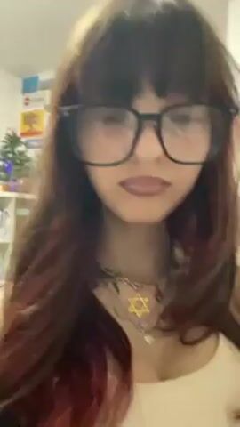 A young woman is centered in the frame, wearing large black glasses and a light-colored tank top. She has long, dark hair with reddish streaks. Around her neck is a necklace featuring a Star of David and other charms. She opens her mouth wide and appears to be singing along to loud music. In the background, slightly out of focus, is a shelf with various items, including what appears to be a small Christmas tree and signs. The room seems to be indoors and well-lit.