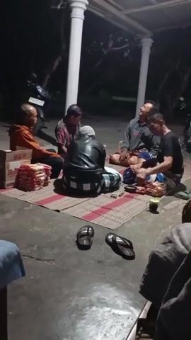 A group of five men are seated on a mat on the ground outside at night. They are gathered under a covered porch supported by white pillars. In the foreground, there are two pairs of flip-flops. To the left, there are two boxes stacked on top of each other, filled with packaged goods. One man in an orange jacket is on the far left, facing the group. Next to him, a man in a red and black plaid shirt is facing the man in orange. In the center, a man wearing a black leather jacket and a white and black checkered sarong faces away from the camera, his head bowed. To his right, a man in a grey sweatshirt is looking towards the man in the black jacket. On the far right, a man in a black t-shirt and patterned sarong is leaning forward, appearing to speak or gesture. A baby is lying on the mat between the man in the grey sweatshirt and the man in the black t-shirt. The ground is concrete. The background is dark with some foliage visible. A motorcycle is partially visible on the left side of the frame.