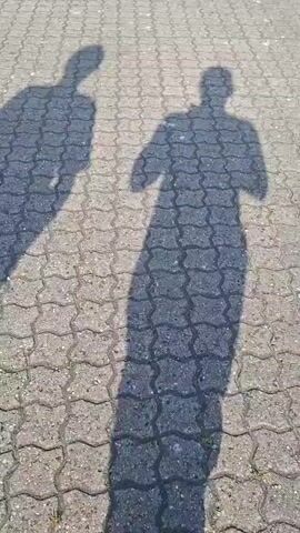 The video shows the shadows of two people on a paved surface. The shadows suggest the people are walking. The pavement is made of interlocking blocks. The light source is bright, indicating a sunny day.