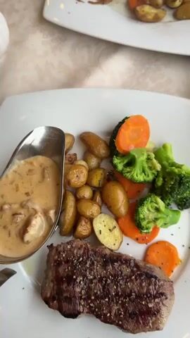 The video begins with a close-up of a plate of food, featuring a steak, roasted potatoes, broccoli, carrots, and a creamy sauce. The plate is white and sits on a table covered with a light-colored tablecloth. In the background, another plate of food with potatoes and what appears to be caramelized onions is visible.

The camera then pans to the right, showing a basket with a slice of flatbread or focaccia. Further to the right, a blurry image of a salad on a white plate with cutlery can be seen, along with a wooden table and chairs. A person's hand wearing a ring is briefly visible, reaching for food.

The video then shifts to a wider shot of the same meal, with another plate of food including steak, potatoes, and vegetables. The surrounding environment is still a restaurant setting, with wooden elements visible.

Finally, the camera pans to the left, revealing a scenic view from what appears to be a balcony or window. Rolling hills, trees, and a distant town are visible under a cloudy sky. A potted plant with purple flowers hangs in the foreground, adding a touch of nature to the scene. The video ends with a blurry view of the landscape.