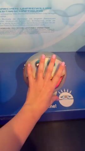 A close-up, medium-shot video shows a person's hand with manicured nails, reaching out and touching a display that features a round, metallic object with a painted face. The face has blue eyes, rosy cheeks, and an exaggerated red mouth with its tongue sticking out. Text on a blue surface below the object reads "Touch me!". The object is mounted on a silver stand. In the background, a blurred panel with text in German and English is visible. The video appears to be taken at an exhibition or museum. The person's hand is blurred as it moves, indicating motion. The camera angle is slightly downward, looking at the display.
