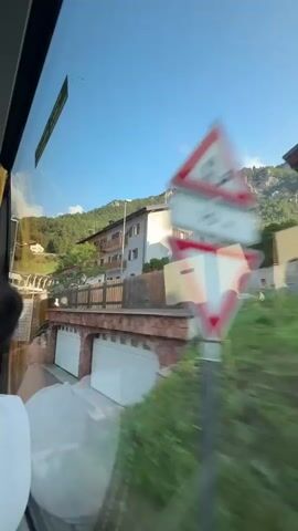 The video shows a view from inside a vehicle, looking out a window. The scene outside is a street in a town, with buildings and a hillside. A white and yellow multi-story building is prominently featured on the right side of the frame. In the foreground, there are two garage doors, one white and one white with a darker stripe. A stone wall runs between the garages and the building above. A signpost with multiple arrows pointing in different directions is visible on the left side of the frame. Two cars are parked on the street in front of the building. The sky is clear and blue.