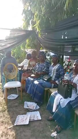 The video shows a group of women sitting under a temporary shelter. The shelter is constructed from dark fabric suspended above them. The women are dressed in a variety of colorful outfits, with several wearing matching blue and white patterned outfits. Some of the women have head coverings. They are sitting in chairs on a grassy surface. Empty plates are on the ground near the chairs, suggesting they have finished eating. The environment appears to be outdoors, possibly a garden or backyard, with trees visible in the background. The sun is shining brightly, creating shadows in the scene.