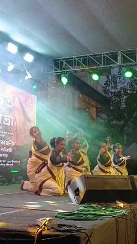 This is a video of a group of young girls performing a traditional dance on a stage. They are dressed in matching white and beige costumes with dark accents. The stage is lit with bright lights, some of which are green, and there is a backdrop with text in an unknown language. The girls are positioned in a line, with the leftmost dancer kneeling and the others standing or in a similar kneeling position, all performing synchronized movements with their arms and hands. The performance appears to be taking place outdoors, possibly in the evening, as indicated by the darkening sky in the background. There are speakers and stage equipment visible in the foreground.
