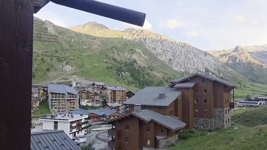 The video captures a panning shot of a mountain village during daytime. The foreground features wooden buildings with stone foundations and pitched roofs, characteristic of alpine architecture. Some buildings have balconies with railings. In the midground, a cluster of similar wooden buildings is visible, along with some modern white apartment blocks. The background is dominated by lush green mountainsides dotted with some sparse vegetation and rocky outcrops. Higher up, snow-capped peaks and glaciers are visible under a bright, partly cloudy sky. A dark, cylindrical object, possibly a pipe or railing, is partially visible in the upper left corner of the frame. The overall impression is a scenic view of a mountain resort, likely during the summer or early autumn season, given the green landscape.