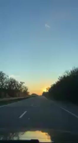 The image captures a perspective from inside a vehicle traveling on a two-lane asphalt road during either sunrise or sunset. The sun, a bright orange orb, is visible low on the horizon directly ahead, casting a warm, brilliant glow that creates a sunburst effect. The sky above transitions from a clear, deep blue at the top to vibrant orange and yellow hues closer to the horizon.

Dense trees and some bare vegetation line both sides of the road, forming a natural corridor. The road itself features clear white dashed lane markings. The setting appears to be a rural or semi-rural highway.

The time of day is the golden hour, characterized by the low-angle, warm sunlight. The weather is clear with no prominent clouds. The scene conveys a sense of peaceful travel and natural beauty. Based on the provided context, the location is Korotych, Ukraine.

No people are visible in the frame, and there is no discernible text within the image. The focus is entirely on the road ahead and the dramatic sky.