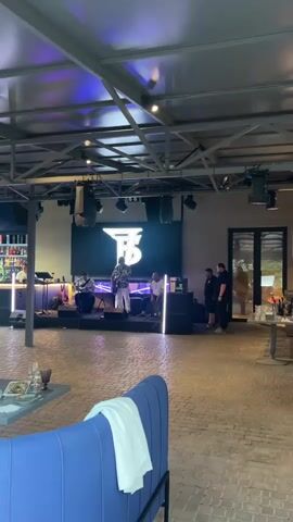 A wide shot captures a dimly lit indoor venue, possibly a restaurant or bar, with a live band performing on a raised stage. The stage backdrop features a large, illuminated logo that appears to be "TB" in a stylized font. Onstage, a male singer, dressed in a patterned shirt and white pants, is actively singing and gesturing. To his left, a male musician plays a keyboard, and to his right, another musician, possibly a drummer, is partially visible. Two other individuals, likely audience members or staff, stand near the stage to the right, observing the performance. The venue has a rustic feel with exposed ceiling beams and brick-like flooring. In the foreground, a plush blue sofa with a white towel draped over it suggests a seating area. To the left, a bar with bottles and glassware is visible. The overall ambiance is relaxed and casual, with subtle purple lighting adding to the atmosphere.