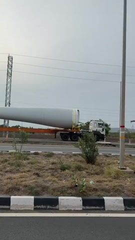 A truck is seen transporting a very long, cylindrical object, which appears to be a wind turbine blade, on a highway. The truck is white and is pulling a flatbed trailer. The blade is gray and extends far beyond the truck and trailer. The scene is outdoors, with a cloudy sky and some vegetation visible along the side of the road. Power lines and a tall transmission tower are also present in the background. The camera appears to be mounted on a moving vehicle, as the perspective shifts slightly throughout the video.