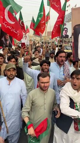 A crowd of men are gathered on a street, holding flags and portraits. The flags are red and green with a crescent moon and star, and some also have the logo PTI. Several portraits show a man's face. The men are wearing traditional clothing such as kurtas and shalwar kameez. Some are shouting and raising their hands. The background shows buildings and a street with some vehicles. The overall impression is of a political rally or demonstration.