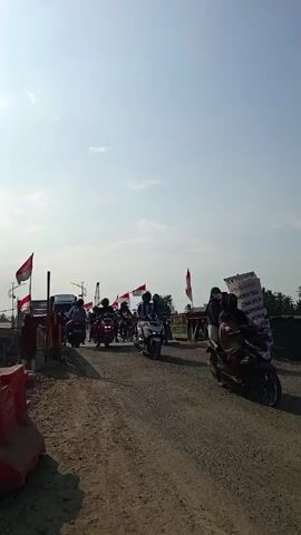 A procession of motorbikes moves across a bridge in the daytime. Indonesian flags are placed intermittently along the bridge. A sign sits on the right side of the bridge. People are seated on the motorbikes. The road surface is rough and unpaved. The sky is light blue with a few clouds.