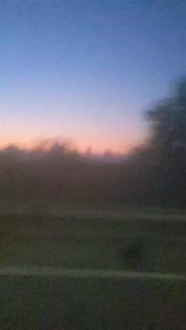 The video shows a blurry landscape at dusk. The sky is a gradient of blue to purple, with hints of pink and orange near the horizon. Clouds are visible, some dark and wispy, others lighter and more defined. Below the sky, a dark, silhouetted line of trees or hills stretches across the frame. In the foreground, there are horizontal bands of lighter and darker green, possibly representing fields or grassy areas, with a white line bisecting the lower half of the image, suggestive of a road. The overall impression is one of movement, as if the footage was captured from a vehicle traveling at speed. There are no people or discernible objects in sharp focus.