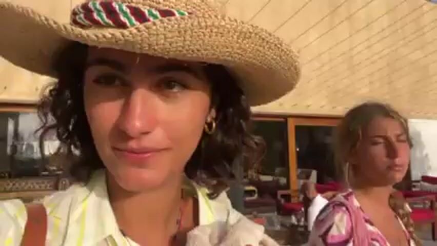 The video shows a young woman in a straw hat holding a tan towel and smiling at the camera. She is standing outside a building with a light beige exterior. In the background, people are sitting on red chairs on a stone patio. The woman then covers her face with the towel and walks away from the camera. Another woman wearing a baseball cap joins her, and they walk together. A man in a white shirt can be seen tending to something on the patio. The scene is outdoors on a sunny day.