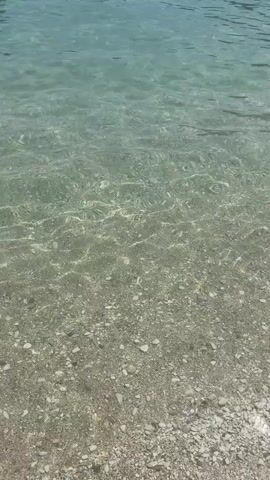 The video shows a clear, shallow body of water with a pebble-strewn bottom. The sunlight glints off the surface of the water, creating shimmering patterns. In the distance, at the upper right of the frame, a few figures can be seen partially submerged in the water, suggesting they are swimming or wading. The overall impression is one of a serene and tranquil aquatic environment, likely a beach or a lake.