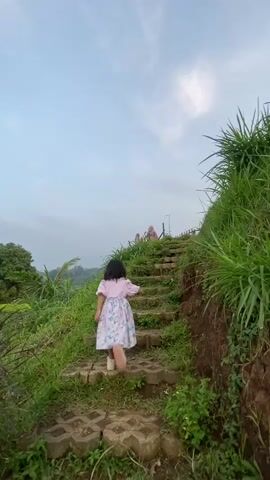The video shows a young girl running up a set of stone steps outdoors. The steps are built into a grassy hillside, with tall grass growing on either side. The girl is wearing a pink top and a floral skirt. She is running towards the top of the steps, where two people can be seen standing on a small platform with a wooden railing. The sky is blue with some clouds. The girl is moving quickly and looks like she is having fun.