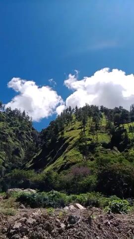 The video shows a landscape view of a mountainous area under a bright blue sky with scattered white clouds. The slopes are covered in lush green vegetation, with trees dotting the hillsides. In the foreground, there is a rocky and debris-strewn area, possibly indicating a landslide or construction site. The camera appears to be handheld and pans slightly, capturing the natural beauty of the environment. There are no people or animals visible in the video. The lighting suggests it is daytime, likely sunny.
