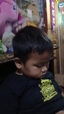 The video shows a young Asian boy with dark hair, wearing a black t-shirt with a yellow design. He is looking down and appears thoughtful, touching his lips with his finger. The background includes various objects, such as toys and furniture, creating a cozy indoor setting. The video is shot at a close range, focusing on the boy's face and upper body.