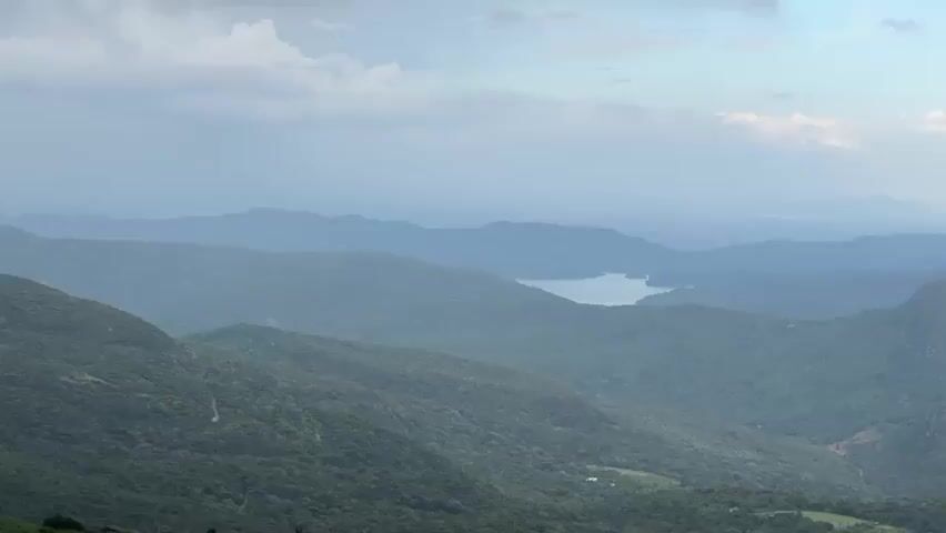 The video shows a panoramic view of a mountainous landscape. Lush green hills and valleys are visible, with a body of water appearing in the distance. The sky is overcast with dark clouds, although some patches of blue are visible. The foreground consists of a grassy hilltop. There are no people or animals visible in the scene. The camera appears to be stationary, capturing the scenery.