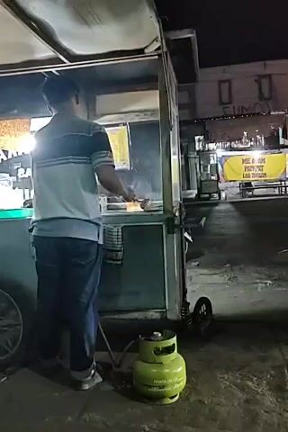 The video shows a man standing behind a food cart at night. The food cart has a propane tank connected to it. The man is wearing a striped shirt and jeans. He appears to be preparing food. In the background, there are other food carts, motorcycles, and buildings. The street is wet, suggesting it may have recently rained. There are lights illuminating the scene.