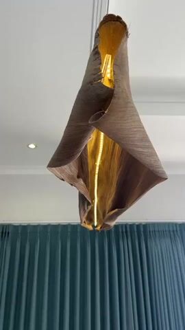 A close-up shot focuses on a unique pendant light fixture made from a large, dried leaf. The leaf is a warm brown color with visible veins and a rough, textured surface. It is suspended from the ceiling by thin wires. The light source, a long, thin bulb, is visible inside the leaf, emitting a warm, golden glow that shines through the leaf's semi-translucent material. The leaf is artfully shaped, appearing to be folded and draped, creating an organic and flowing form. The background shows a white ceiling with a subtle coving and a recessed light. Below the light fixture, dark teal curtains are visible, creating a contrast with the lighter tones of the fixture and ceiling. The overall impression is of an artistic and natural lighting element in a room.