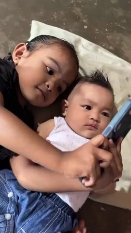 Two young children, a girl and a baby, are lying down. The girl has dark hair in a braid and is wearing a black shirt and blue jeans. She is holding a smartphone in front of the baby, who is wearing a white tank top and blue jeans. The baby is looking at the phone with a neutral expression. The girl is looking at the baby and smiling slightly. They are on a light-colored blanket on a dark surface. The camera is positioned above and slightly to the side of the children.