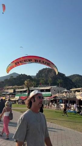 The video shows a paraglider with "DEEP BLUE UFO" written on it, descending towards a beach area. Several people are visible on the beach, some walking and others watching the paraglider. There are buildings and palm trees in the background, suggesting a tourist resort. The paraglider is bright orange and yellow, with red lettering. Two people are assisting the paraglider as it lands, one of whom is wearing a helmet. The ground is paved with stone tiles, and there is a grassy area to the right. The sky is clear and blue.