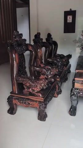 The video pans across a living room area displaying ornate, dark wood furniture. The focus is on a large sofa with intricate carvings, possibly depicting dragons or mythical creatures. To the right of the sofa is a matching coffee table, also elaborately carved. The furniture appears to be made of dark wood with lighter wood accents, possibly ebony or rosewood. The room has light-colored walls and a tiled floor. There are a few decorative items visible, including a vase with dark red decorative branches on the coffee table and a framed calendar or picture on the wall behind the furniture. The overall impression is one of luxury and traditional craftsmanship.