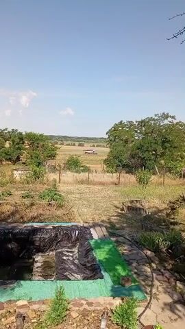 The video shows a natural landscape with a clear blue sky. In the foreground, there is a black tarp covering an area, possibly a pond or pool. To the right of the tarp, there is a strip of green material. Beyond this, the ground is dry and covered in grass and some scattered plants. A wire fence runs across the middle ground. In the distance, there are trees and fields, with a small white structure visible in the far distance. The scene appears to be outdoors on a sunny day.