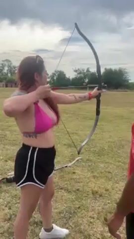A woman with red hair and a pink bikini top, wearing black shorts with white stripes, is practicing archery. She is holding a long black bow, aiming an arrow towards a target in the distance. She has tattoos on her arms and stomach. A man in a red shirt stands to her right, guiding her stance. The scene is set outdoors on a grassy field under a cloudy sky, with some trees in the background.