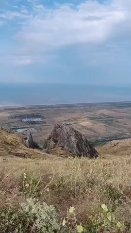 A video pans across a landscape from a high vantage point. In the foreground, dry grass and some small plants grow on a gentle slope. Further down the slope, large rocks are visible. Beyond the rocks, a flat, cultivated landscape stretches into the distance, with some scattered buildings. The sky above is blue with scattered white clouds. The camera pans slowly to the right, revealing more of the distant landscape and the horizon. There are no people or animals visible in the video. The overall impression is one of a rural, somewhat arid environment under a cloudy sky.