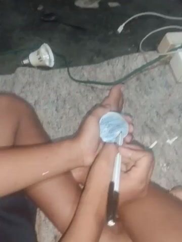 The video shows a person's hands holding a small, circular object, possibly made of paper, in their left palm. The person is using a marker in their right hand to write on the object. The writing appears to be in blue ink. The background is a textured surface, possibly a floor or rug, with a dark green area on the left side. Several objects are visible in the dark green area, including a white power strip with cords, and a light bulb that appears to have writing on it. The scene is dimly lit.