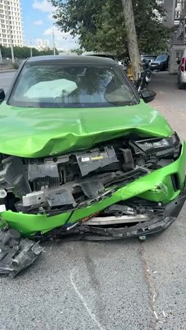 The video shows a severely damaged bright green car, likely after a car accident. The front of the car is caved in, with the hood crumpled and the bumper dislodged. The headlights are broken and hanging loosely, and the radiator and other engine components are exposed and twisted. The airbags on the driver's side have deployed, suggesting the impact was significant. The car is parked on the side of a street, with a sidewalk and some greenery visible in the background. Other vehicles and buildings can be seen in the distance, indicating an urban or suburban setting. The overall scene depicts the aftermath of a collision, with the car appearing to be irreparable.